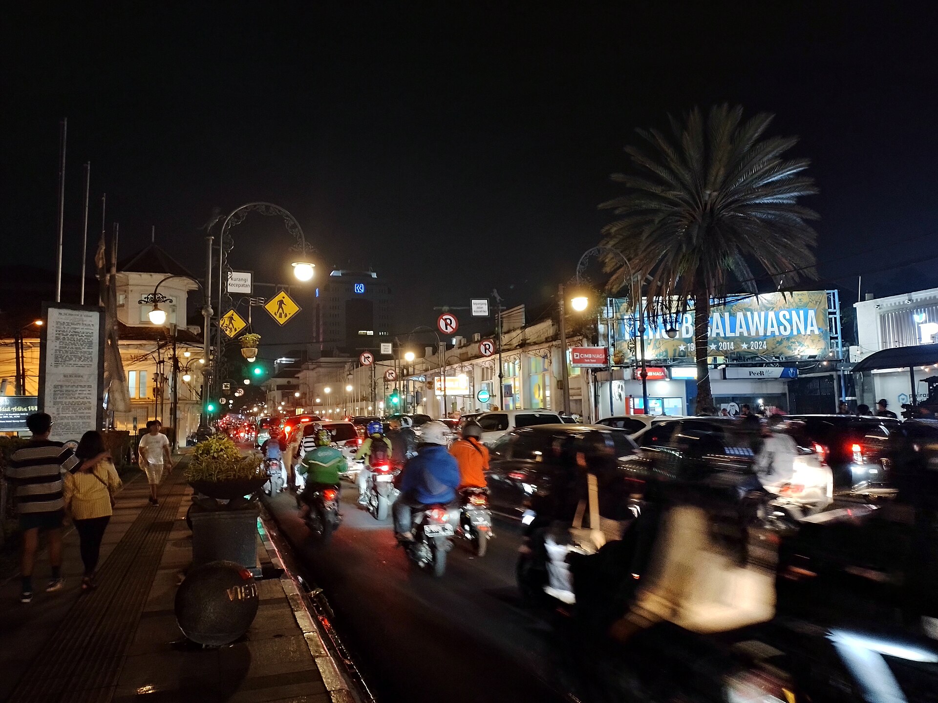 Asia-Afrika street in Bandung at night, lit shopfronts and glowing signs. Photograph by RasyaAbhirama13, CC BY-SA 4.0.